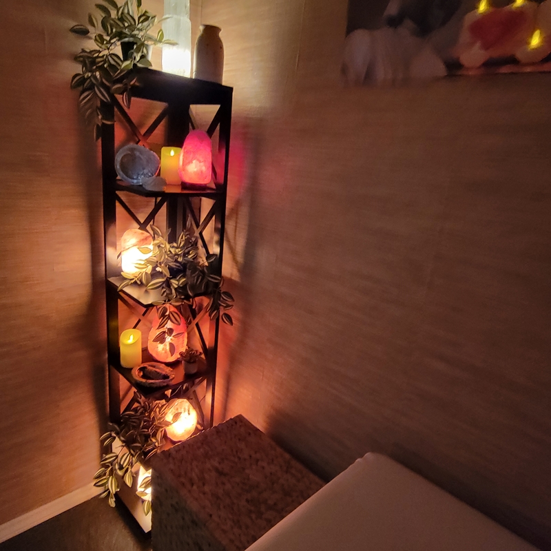 Corner shelf at the foot of the Reiki bed in The Zen Shop Healing Room filled with Himalayan salt lamps, selenite tower, candles, crystals, and greenery