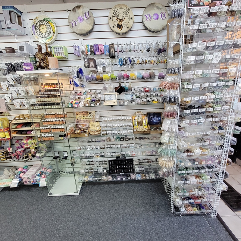 View of the back half of the north wall at The Zen Shop Oshawa, featuring bins of rough crystals and pocket tokens, QR-coded tumbled stones, shaped crystals like hearts and worry stones, essential oils, spiritual keychains, evil eye items, and tribal-style drums mounted above.