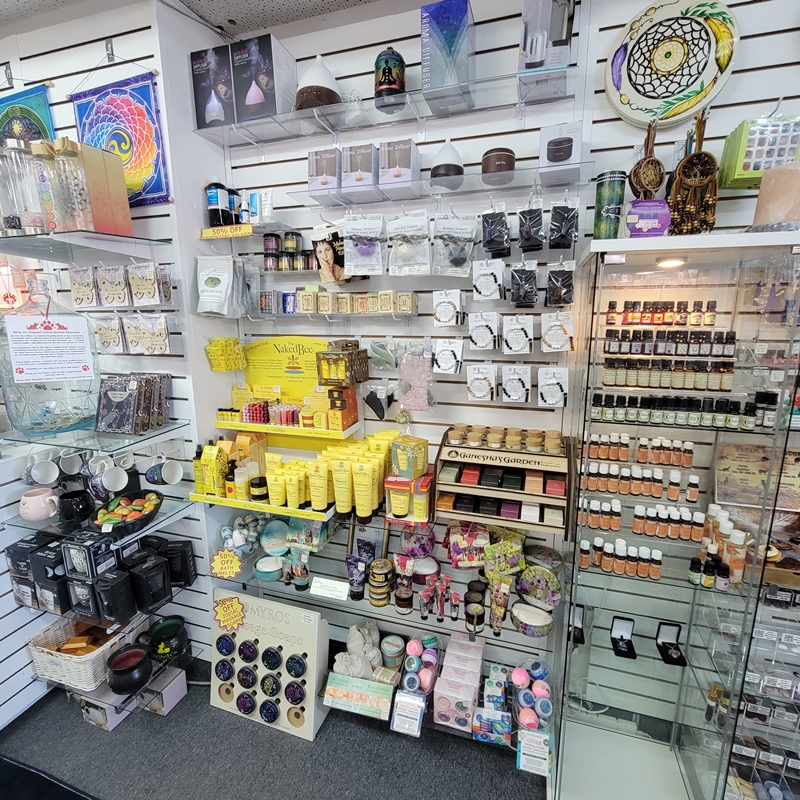 Back north corner of The Zen Shop Oshawa showcasing a variety of self-care products including Naked Bee and Barefoot Venus body care, bath bombs, essential oils, jewellery diffusers, gua shas, crystal eye masks, and spiritual-themed mugs.