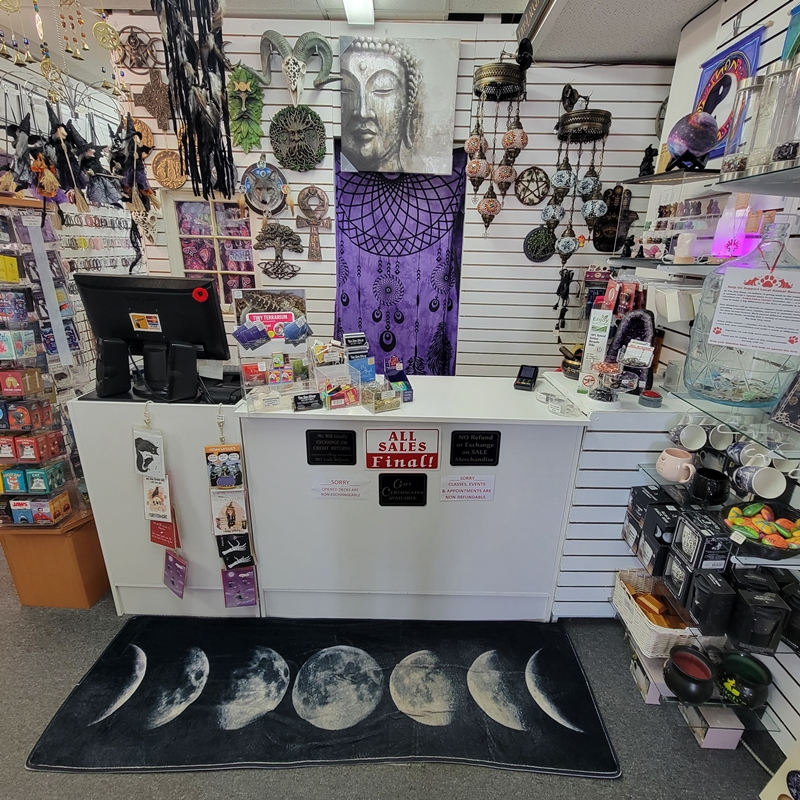 Back-of-store checkout counter at The Zen Shop Oshawa featuring a display of spiritual novelty items, oracle decks, wall plaques, hanging décor, and spiritual mugs. A moon phase rug adds a mystical touch to the customer experience.