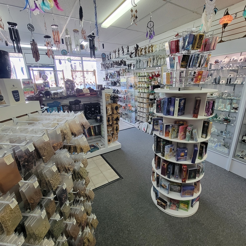Centre aisle view at The Zen Shop Oshawa featuring a rotating oracle card display and racks of dried herbs, with crystal cabinets, metaphysical books, spiritual décor, and hanging wind chimes visible in the background.