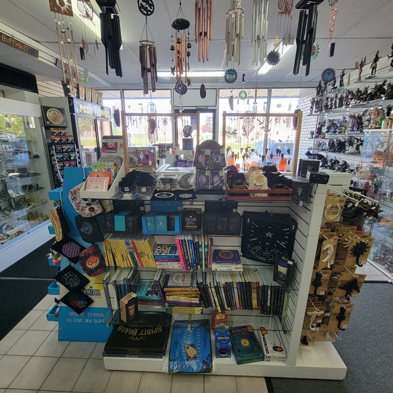 Centre-front view at The Zen Shop Oshawa showcasing pendulums, pendulum mats, metaphysical books, how-to oracle decks, Spirit Boards, Flying Wish Paper, storage pouches, and decorative wall hooks. Hanging wind chimes and crystal figures line the surrounding walls.