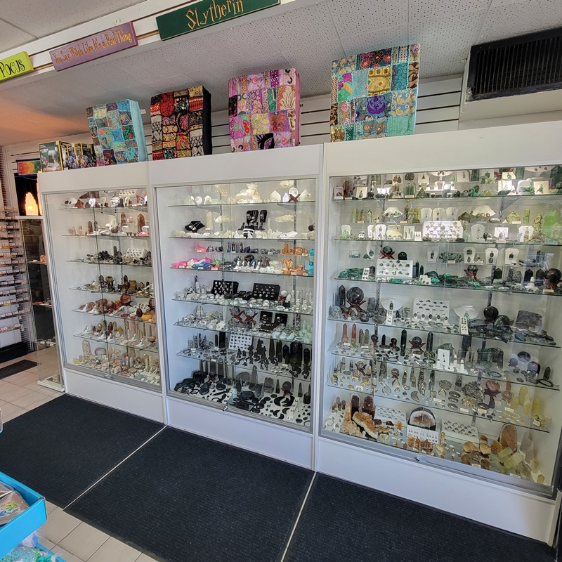 Crystal display cabinets along the north wall of The Zen Shop Oshawa, filled with carved gemstones, crystal spheres, towers, and jewellery, with colorful meditation cushions stacked above.