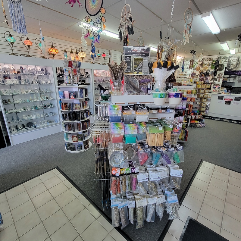 Mid-store view at The Zen Shop Oshawa looking toward the centre and south wall, featuring smudging herbs and accessories, hanging dream catchers, oracle and tarot cards, and crystal jewellery cabinets.
